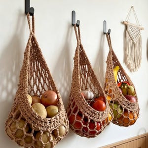 Peut inclure: Trois paniers suspendus en macramé beige accrochés à des crochets muraux noirs. Les paniers sont fabriqués en corde épaisse avec un motif en filet ouvert et contiennent divers produits frais, notamment des pommes de terre, des oignons, de l'ail, des tomates et des bananes.