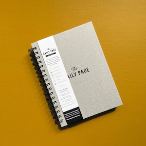 Puede incluir: Un cuaderno gris de espiral con el texto "The Daily Page" en la portada. El cuaderno tiene un diseño en blanco y negro con una lista de hábitos diarios para llevar un seguimiento, incluyendo hidratación, nutrición, movimiento, comunidad, sueño y salud mental. El cuaderno está sobre una superficie amarilla.