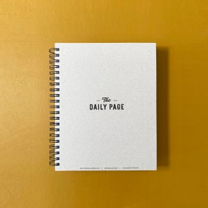 Puede incluir: Una libreta de espiral blanca con el texto "The Daily Page" impreso en negro en la portada.