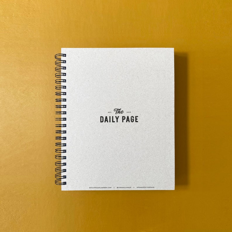 Puede incluir: Una libreta de espiral blanca con el texto "The Daily Page" impreso en negro en la portada.