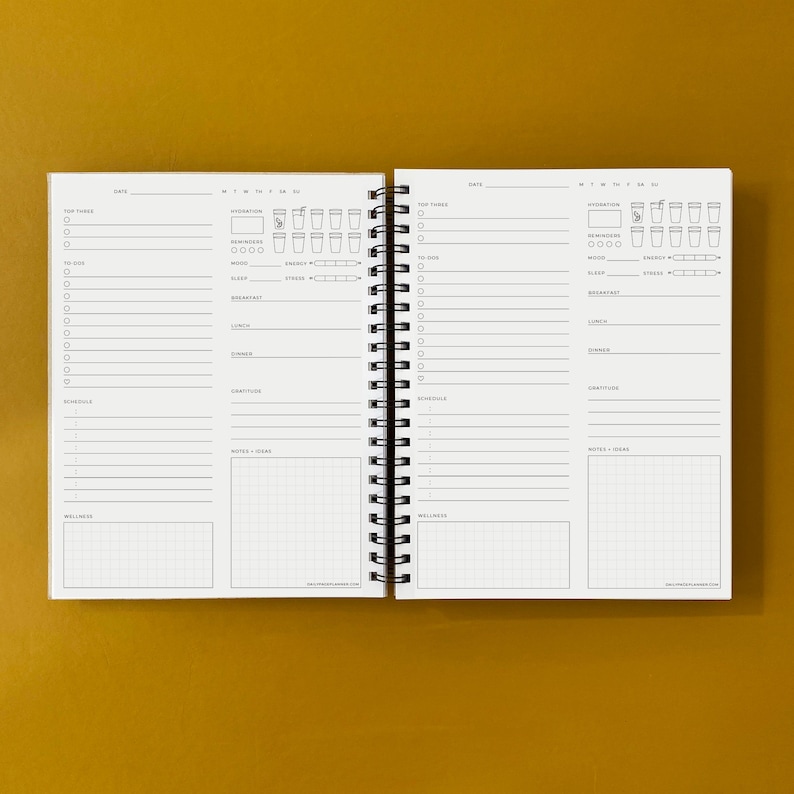 Puede incluir: Una agenda blanca con una encuadernaci&oacute;n en espiral negra. La agenda tiene secciones para tareas diarias, seguimiento de la hidrataci&oacute;n, estado de &aacute;nimo, sue&ntilde;o, energ&iacute;a, estr&eacute;s, notas y bienestar. La agenda est&aacute; abierta en dos p&aacute;ginas, ambas con el mismo dise&ntilde;o.