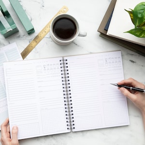 Puede incluir: Un planificador de espiral abierto con fondo blanco y l&iacute;neas negras. El planificador tiene una secci&oacute;n para tareas diarias, una secci&oacute;n para la planificaci&oacute;n de comidas y una secci&oacute;n para la ingesta de agua. Una mano sostiene un bol&iacute;grafo y escribe en el planificador.