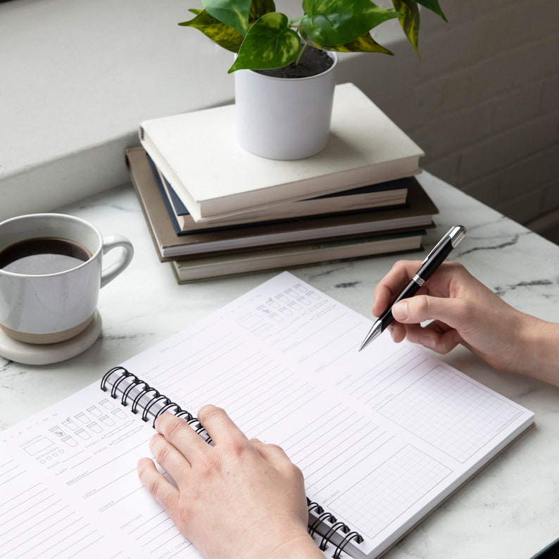 Puede incluir: Un cuaderno blanco de espiral con un bol&iacute;grafo negro sobre una superficie de m&aacute;rmol blanco. El cuaderno tiene un patr&oacute;n de cuadr&iacute;cula y l&iacute;neas para escribir. Una mano sostiene el bol&iacute;grafo y escribe en el cuaderno.