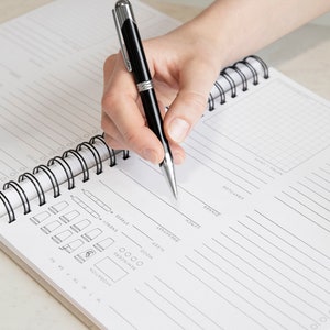 Puede incluir: Un cuaderno blanco de espiral con l&iacute;neas negras y un patr&oacute;n de cuadr&iacute;cula. El cuaderno tiene secciones para entradas diarias, incluyendo fecha, hidrataci&oacute;n, recordatorios, estado de &aacute;nimo, energ&iacute;a, sue&ntilde;o, estr&eacute;s, desayuno, almuerzo, cena, gratitud y notas. Una mano sostiene una pluma negra y escribe en el cuaderno.