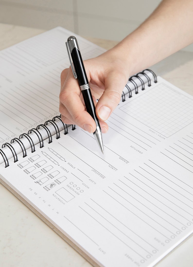 Puede incluir: Un cuaderno blanco de espiral con l&iacute;neas negras y un patr&oacute;n de cuadr&iacute;cula. El cuaderno tiene secciones para entradas diarias, incluyendo fecha, hidrataci&oacute;n, recordatorios, estado de &aacute;nimo, energ&iacute;a, sue&ntilde;o, estr&eacute;s, desayuno, almuerzo, cena, gratitud y notas. Una mano sostiene una pluma negra y escribe en el cuaderno.
