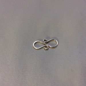 Puede incluir: Cierre de metal plateado con un dise&ntilde;o de ocho. El cierre es un hallazgo de joyer&iacute;a utilizado para conectar cadenas o cordones.