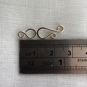 May include: Silver-toned metal clasp with a hook and loop design, measuring approximately 1 inch in length. The clasp is shown next to a ruler for size reference.