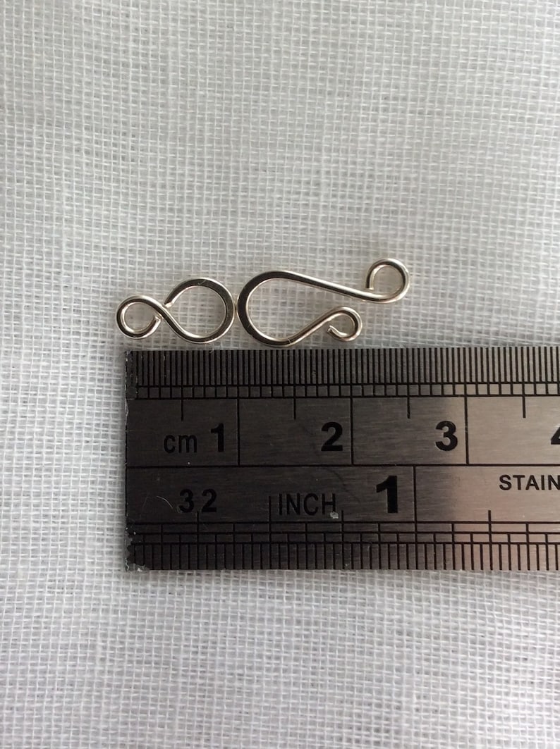 May include: Silver-toned metal clasp with a hook and loop design, measuring approximately 1 inch in length. The clasp is shown next to a ruler for size reference.