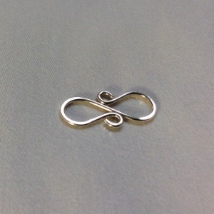 Puede incluir: Un cierre de metal plateado con un dise&ntilde;o de ocho. El cierre es un hallazgo de joyer&iacute;a utilizado para conectar cadenas o cordones.