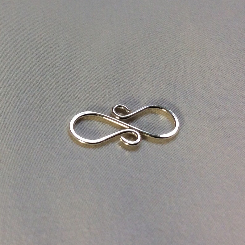 Puede incluir: Un cierre de metal plateado con un dise&ntilde;o de ocho. El cierre es un hallazgo de joyer&iacute;a utilizado para conectar cadenas o cordones.