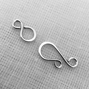 May include: Two silver metal clasps for jewelry making. One clasp is a simple loop, the other is a more elaborate, curved design.