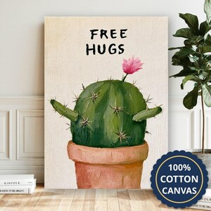 Op de afbeelding: Een canvas print met een groene cactus in een terracotta pot met de tekst FREE HUGS in het zwart erboven. De cactus heeft een enkele roze bloem aan de bovenkant en twee uitgestrekte armen, geschilderd met zichtbare penseelstreken.
