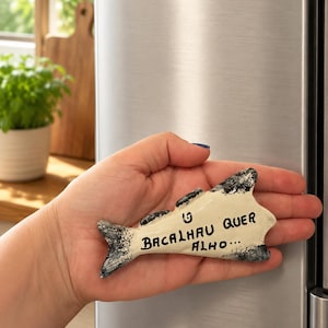 Könnte beinhalten: Ein fischförmiger Magnet mit cremefarbenem Körper und dunkelgrauen, texturierten Rändern. Der Text "BACALHAU QUER ALHO..." ist in Schwarz in der Mitte des Fisches geschrieben. Der Magnet ist für die Anbringung auf Metalloberflächen vorgesehen.