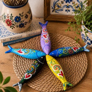 Pode incluir: Cinco figuras de peixes em madeira pintadas à mão dispostas em forma de estrela. Cada peixe apresenta uma cor única—roxo, azul, azul escuro, preto e amarelo—e é decorado com padrões complexos de pontos coloridos e um motivo de coração.