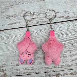 May include: Two pink plush bat keychains. One keychain shows the bat's face with purple ears and a smiling expression. The other keychain is a solid pink bat shape. Both keychains have a silver ring and pink ribbon.