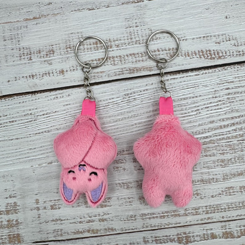May include: Two pink plush bat keychains. One keychain shows the bat's face with purple ears and a smiling expression. The other keychain is a solid pink bat shape. Both keychains have a silver ring and pink ribbon.
