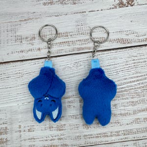 May include: Two plush blue bat keychains. One bat is shown upside down, with white embroidered details for the eyes and mouth. The other keychain shows the back of the bat. Both keychains have a silver ring and a blue ribbon.
