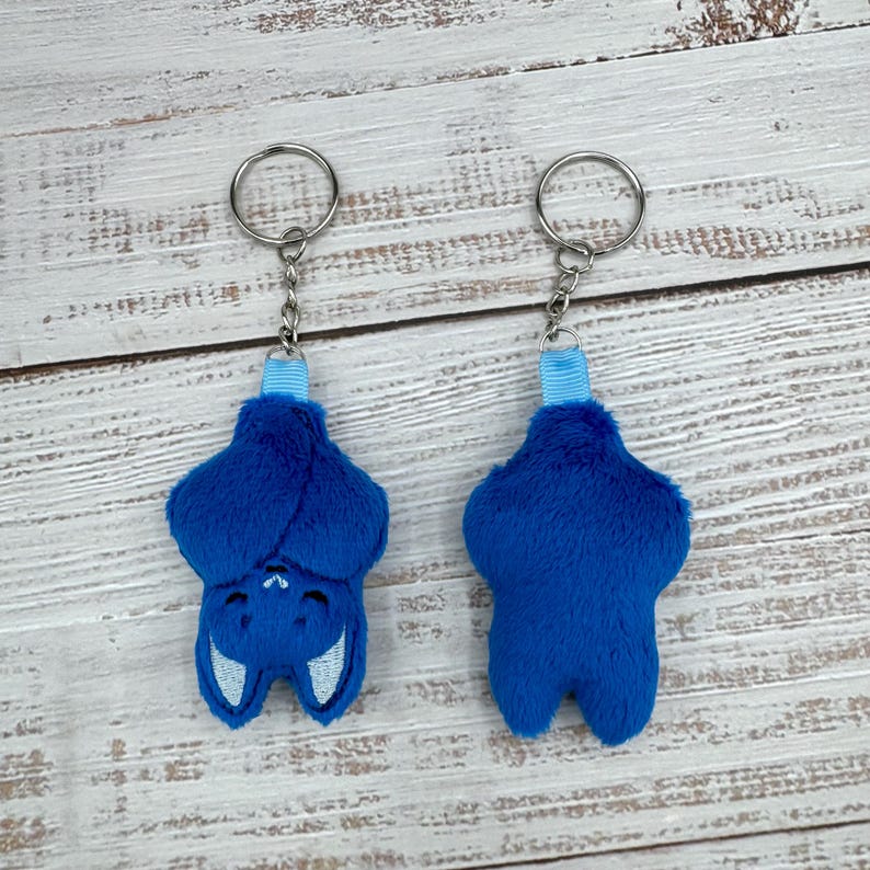 May include: Two plush blue bat keychains. One bat is shown upside down, with white embroidered details for the eyes and mouth. The other keychain shows the back of the bat. Both keychains have a silver ring and a blue ribbon.