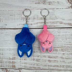 May include: Two plush bat keychains. One is blue with light blue accents, the other is pink with purple accents. Both have silver key rings and matching colored ribbon loops.