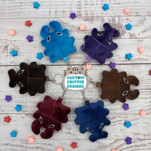 May include: A collection of plush keychains in various colors, including blue, purple, brown, and burgundy. Each keychain features a unique creature design with embroidered details and a metal keyring. The keychains are arranged around a central charm that reads "CUSTOM CRITTER DESIGNS."