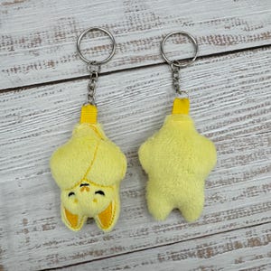 May include: Two yellow plush bat keychains. Each keychain features a fluffy, star-shaped body with a yellow loop and silver chain. One bat has embroidered facial features and ears. The other keychain shows the back of the bat.