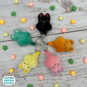 May include: A collection of plush bat keychains in various colors: black, green, orange, yellow, and pink. Each bat has embroidered details and is attached to a silver keyring. The background includes star-shaped decorations and the text "Custom Critter Designs".