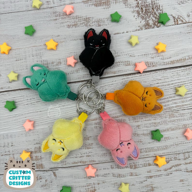 May include: A collection of plush bat keychains in various colors: black, green, orange, yellow, and pink. Each bat has embroidered details and is attached to a silver keyring. The background includes star-shaped decorations and the text "Custom Critter Designs".