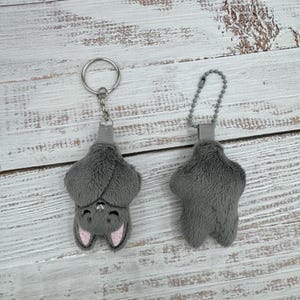 May include: Two gray plush bat keychains. One bat is shown from the front with pink ears and a smiling face. The other bat is shown from the back. Both have a gray ribbon and a metal chain or ring.