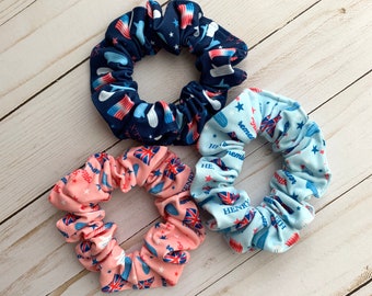 Può includere: Tre scrunchies in tessuto con diversi motivi. Il primo scrunchie è blu con stelle bianche e strisce rosse. Il secondo scrunchie è azzurro con stelle bianche e testo rosso. Il terzo scrunchie è rosa con stelle bianche e testo rosso.