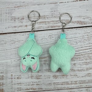 May include: Two mint green bat-shaped keychains. One keychain shows the front of the bat with embroidered pink ears and a smiling face. The other keychain shows the back of the bat. Both keychains have a silver chain and a teal ribbon loop.