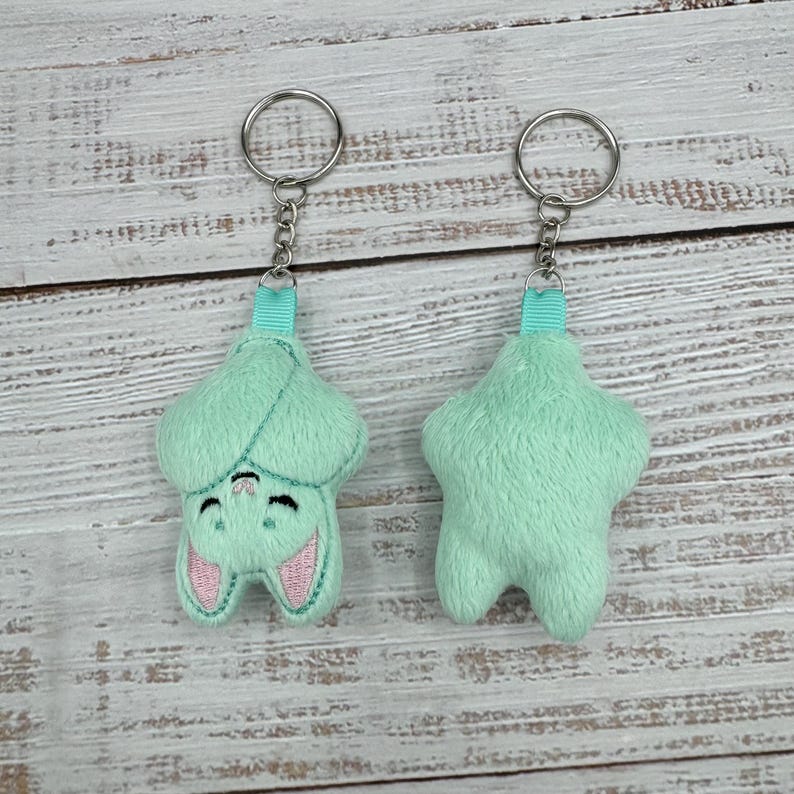 May include: Two mint green bat-shaped keychains. One keychain shows the front of the bat with embroidered pink ears and a smiling face. The other keychain shows the back of the bat. Both keychains have a silver chain and a teal ribbon loop.