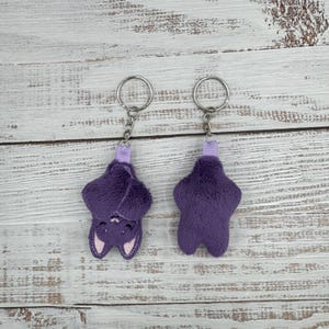 May include: Two plush purple bat keychains with silver key rings. One bat is upside down, showing a smiling face with pink accents. The other bat is upright. Both have a small purple ribbon loop.