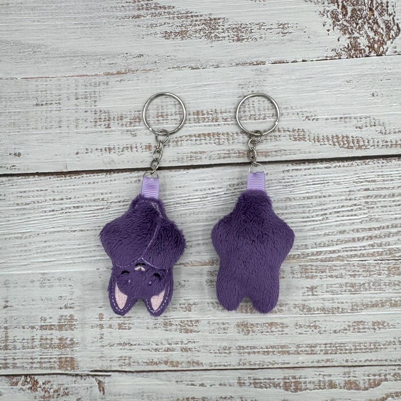 May include: Two plush purple bat keychains with silver key rings. One bat is upside down, showing a smiling face with pink accents. The other bat is upright. Both have a small purple ribbon loop.