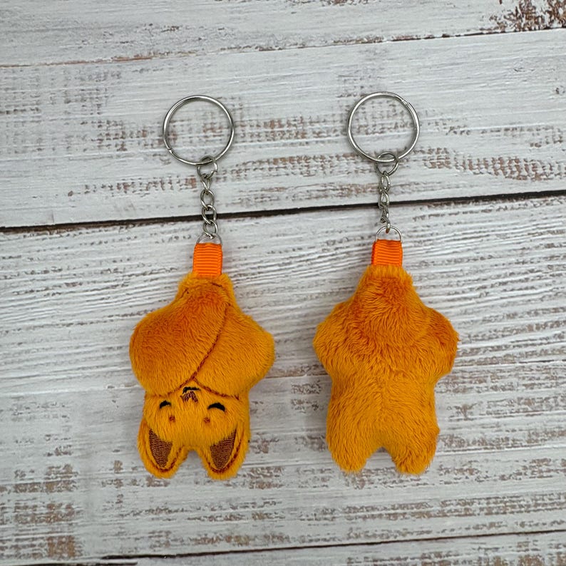May include: Two orange plush bat keychains. One bat is shown from the front with a smiling face and the other from the back. Each keychain has a silver ring and an orange fabric loop.