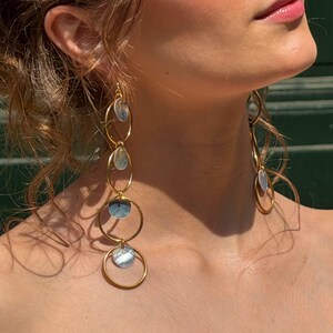 Può includere: Lunghi orecchini pendenti color oro caratterizzati da una serie di quattro cerchi intrecciati. Ogni cerchio contiene un piccolo inserto circolare iridescente in madreperla. Gli orecchini pendono verticalmente.