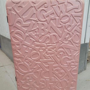 May include: A dusty rose pink hard-shell suitcase featuring an embossed, all-over pattern of various uppercase English alphabet letters. The rectangular luggage has rounded corners and is equipped with black swivel wheels at the base.