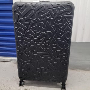 Puede incluir: Una maleta negra vertical con una superficie texturizada que presenta un patrón en relieve de letras mayúsculas superpuestas. El equipaje cuenta con cuatro ruedas multidireccionales en la base y un asa retráctil en la parte superior.
