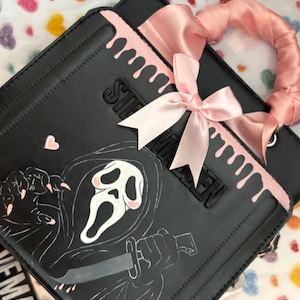 May include: A black square handbag featuring a white ghost-like figure with a knife, pink accents, and a pink satin bow. The bag has a pink drip design at the top, a pink scrunchie-style handle, and the text STEVE MADDEN embossed on the front surface.
