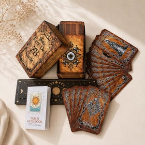 May include: A Gothic Tarot Deck set featuring 78 cards with brown, weathered-look backgrounds and black ink illustrations. The set includes a matching storage box, a black wooden card holder with moon phase icons, and a white Tarot Guidebook.
