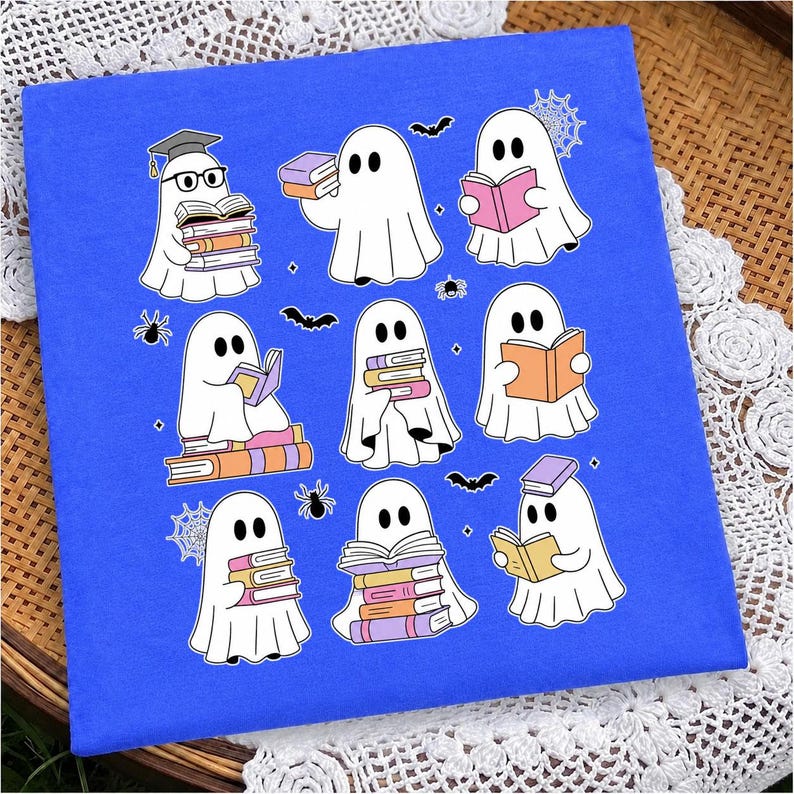 May include: A bright blue t-shirt featuring a graphic print of nine white ghosts reading various colorful books. The ghosts are surrounded by small black spiders, spiderwebs, and bats. One ghost wears a graduation cap and black glasses.