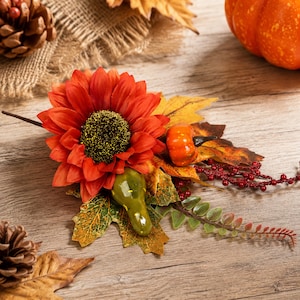 May include: A decorative autumn floral arrangement featuring a large orange sunflower with a dark center, a small glossy orange pumpkin, a green gourd, yellow maple leaves, and a sprig of red berries on a thin brown stem for seasonal display.