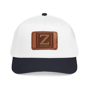 Peut inclure: Une casquette de baseball blanche avec une visière bleu marine foncé et un petit bouton bleu marine foncé sur le dessus. Au centre se trouve un écusson rectangulaire en cuir marron avec une grande lettre Z et le texte ZENTARA gravé.