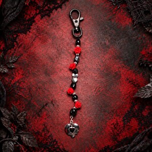May include: A keychain featuring a black metal lobster clasp attached to a strand of alternating black beads, small red rose-shaped charms, and silver-toned heart-shaped beads, ending with a larger silver-toned heart charm at the bottom.