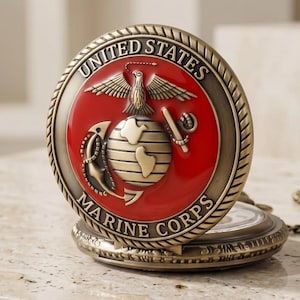 Op de afbeelding: Een rond zakhorloge met een antieke bronzen afwerking, voorzien van het embleem van het United States Marine Corps op het deksel. Het embleem toont een rode cirkelvormige achtergrond met een gouden adelaar, wereldbol en anker, omringd door de tekst UNITED STATES MARINE CORPS.