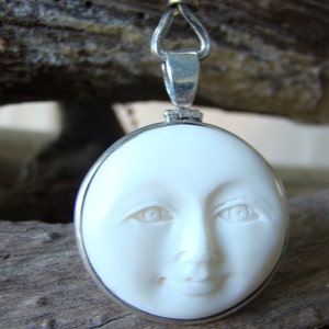 May include: A white carved pendant with a face of a woman. The pendant is oval shaped and has a silver bail.