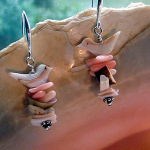 Puede incluir: Un par de pendientes de plata con abalorios de pájaros de concha rosa y blanca. Los pendientes cuelgan de una superficie blanca.