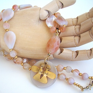 May include: A multi-strand necklace with a pendant featuring two gold angels. The necklace is made with peach and white agate beads and gold spacers.