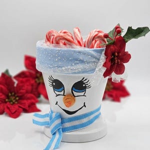 Peut inclure: Un pot en céramique blanc peint avec un visage de bonhomme de neige, doté d'une bordure en maille bleue scintillante, d'une écharpe en tissu rayé bleu et blanc et d'un nez texturé orange. Le pot contient des sucres d'orge et une fleur de poinsettia.