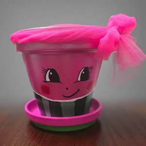 May include: A terracotta plant pot painted in bright pink with a hand-painted cartoon face featuring large eyes, eyelashes, and a smile. The lower section has vertical black stripes. A bright pink tulle fabric bow is tied around the rim.