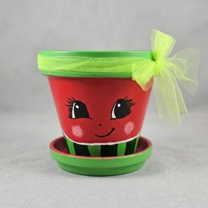 May include: A terracotta plant pot and matching saucer painted to resemble a watermelon with a red center, black seeds, and a green rind. The pot features a hand-painted face with large eyes and pink cheeks, accented by a bright lime green tulle bow.